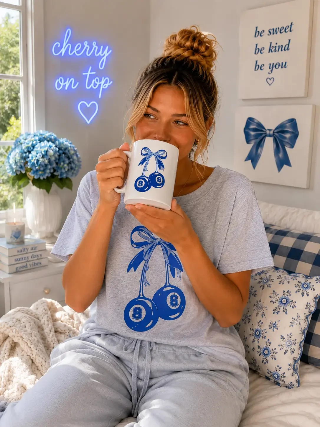 Woman in grey loungewear with a blue eight-ball cherry design drinks from a matching mug in a bedroom.