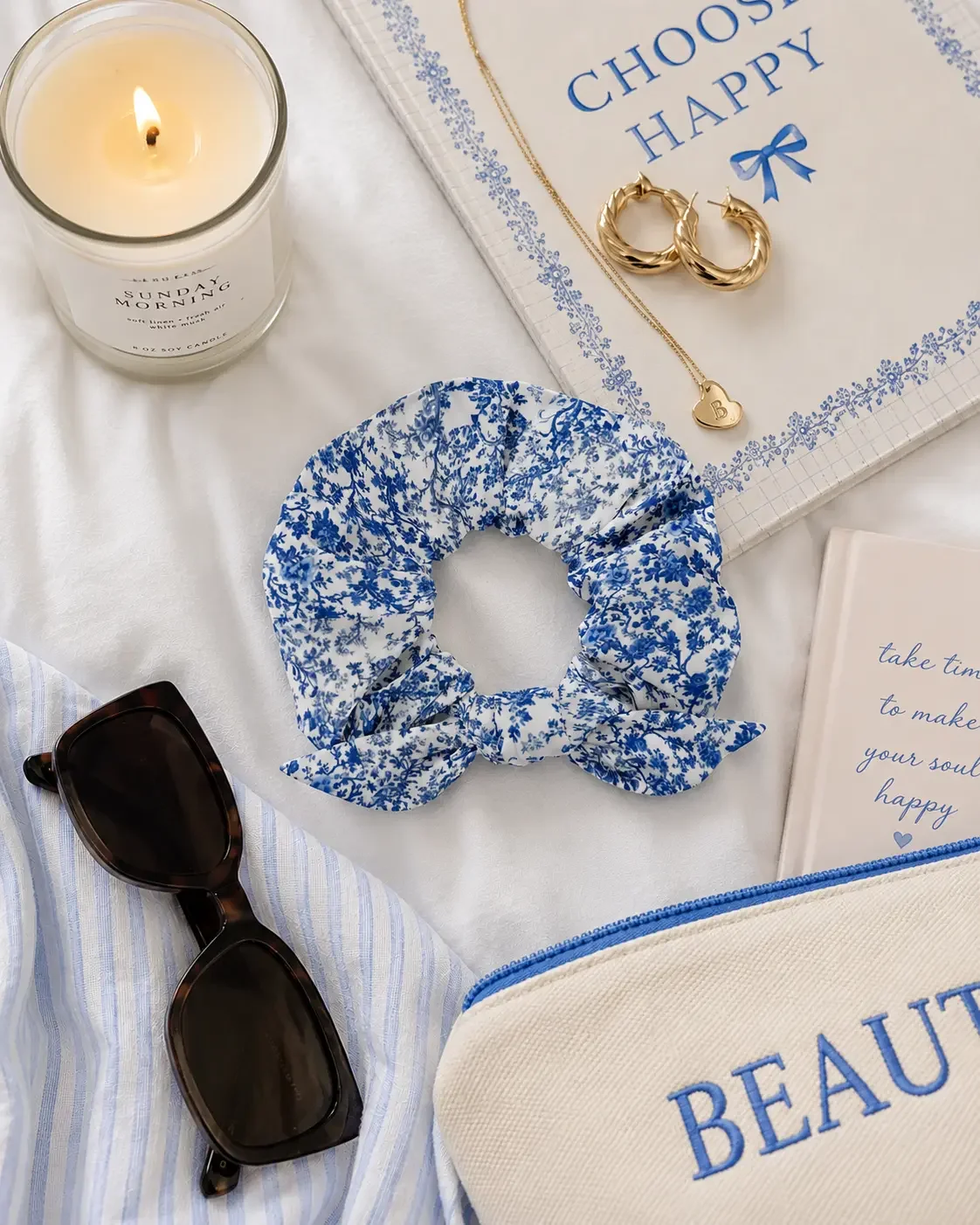 Flatlay of a blue floral bow scrunchie with sunglasses, a candle, books, gold jewelry, and a makeup pouch.