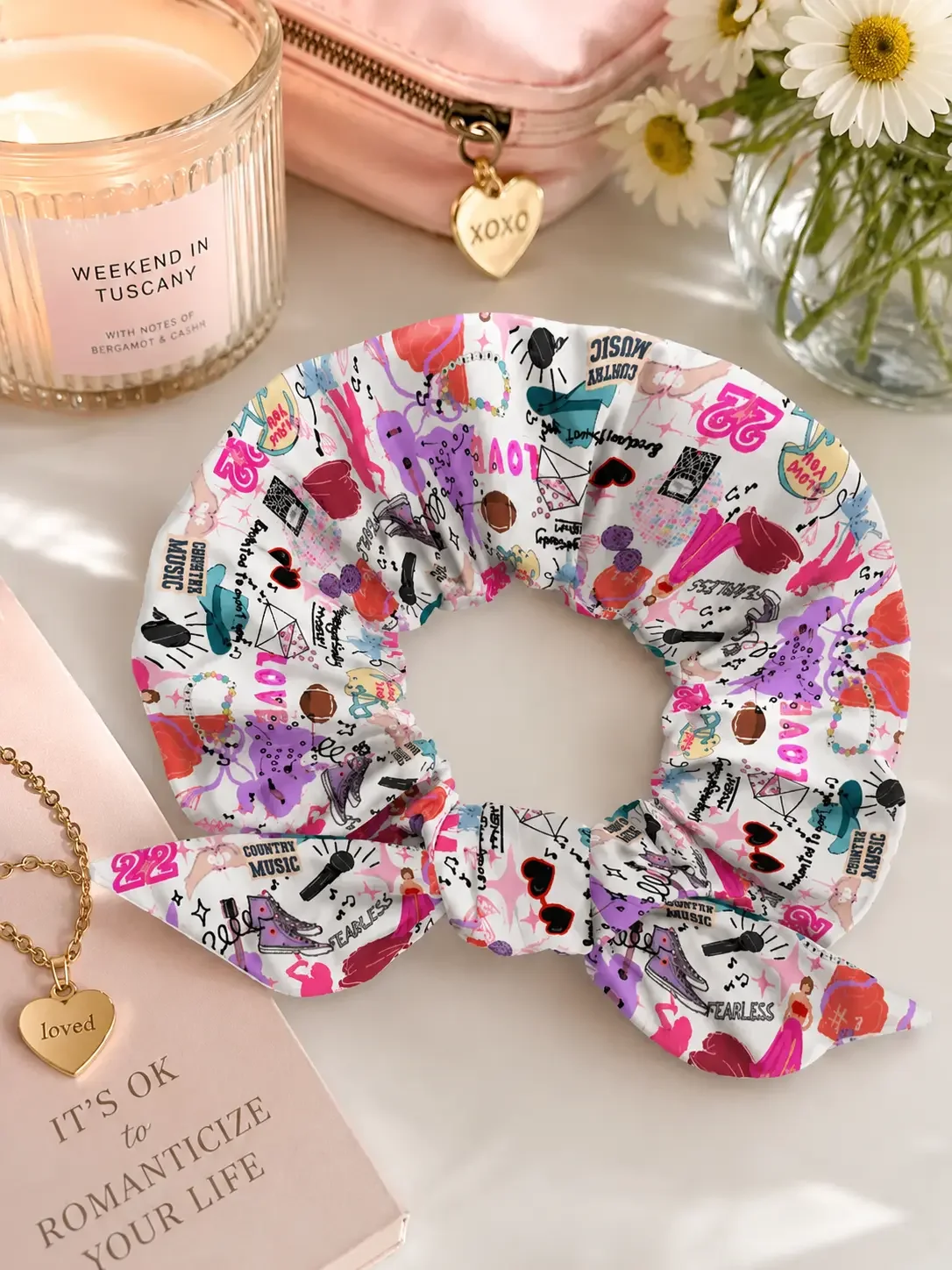 Taylor Swift Eras doodle bow scrunchie displayed flat on a white surface with lifestyle accessories and cosmetics.