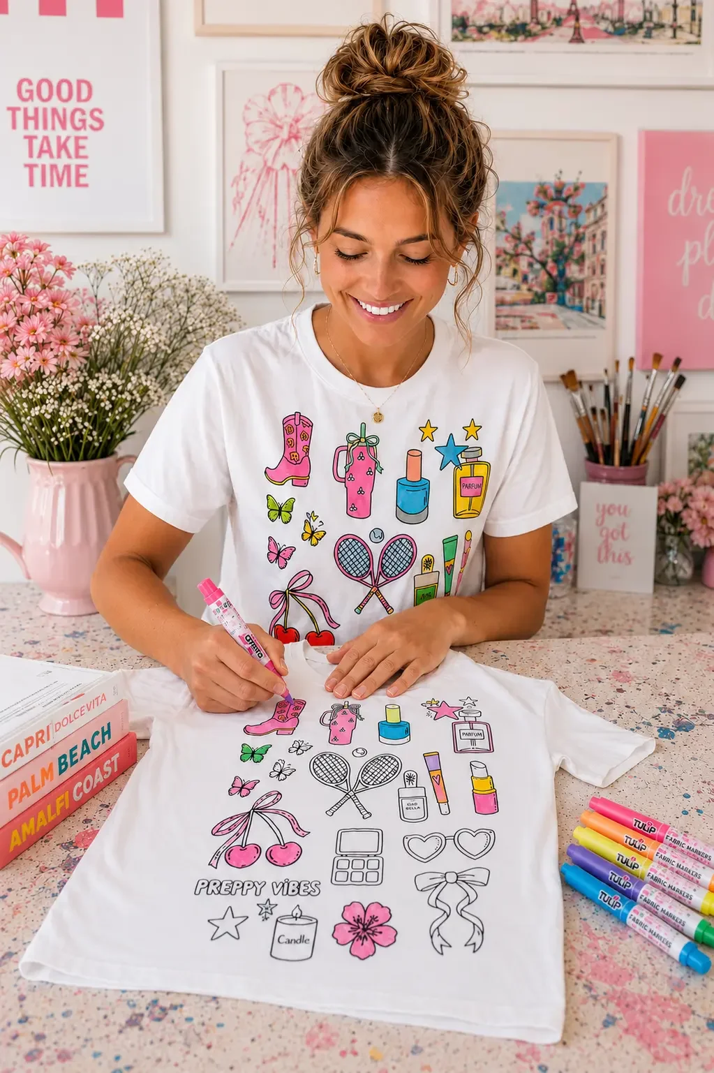 A smiling young brunette woman with a top knot bun hairstyle sitting at a desk and coloring a white graphic t-shirt featuring a preppy vibes doodle design using pink fabric markers.