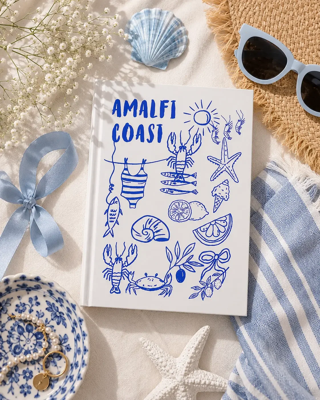Flat lay of a white "Amalfi Coast" journal with blue doodles on a beach towel with a starfish and sunglasses.