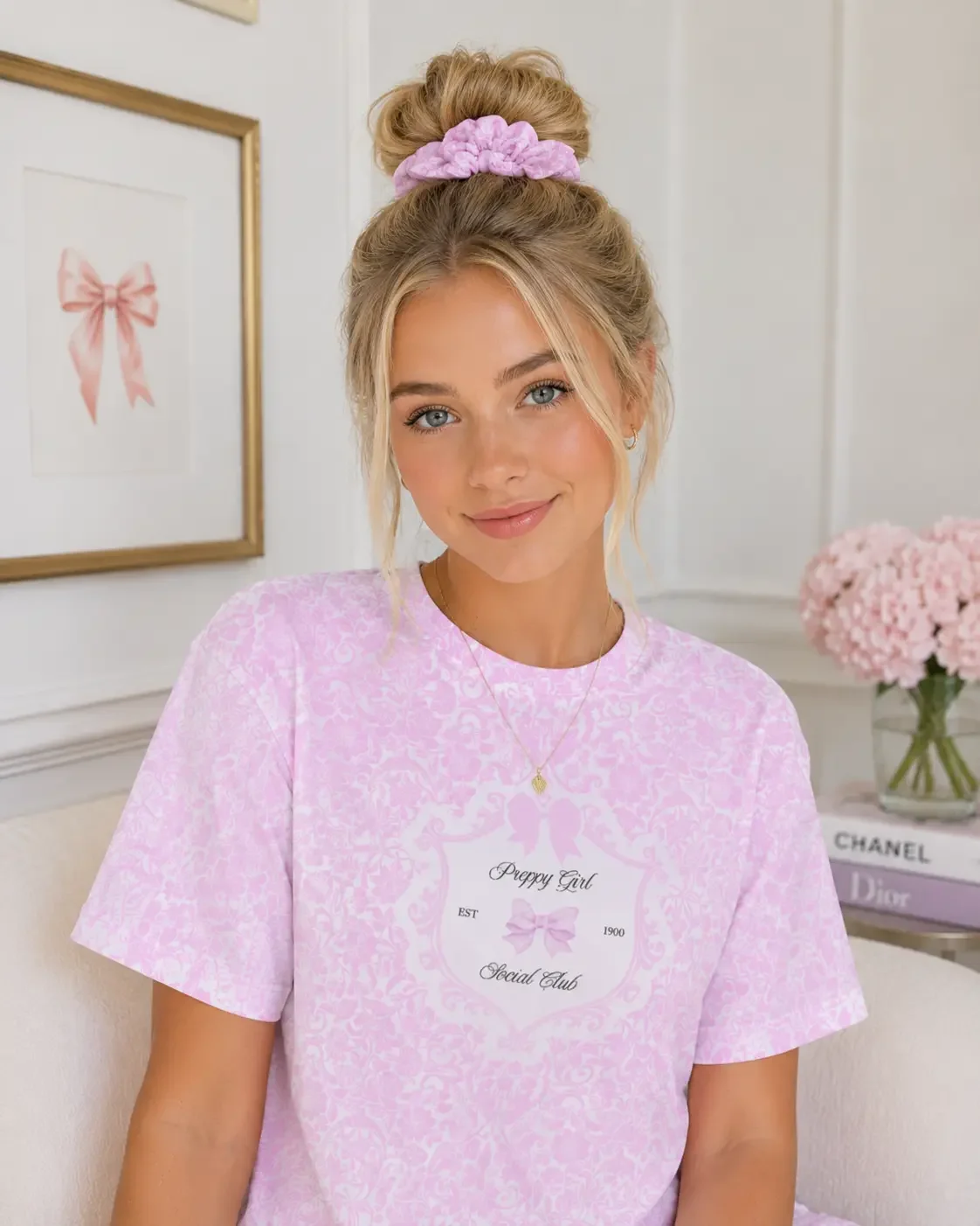 Smiling blonde woman on a white sofa with a high top knot bun, pink patterned scrunchie, and matching graphic t-shirt.