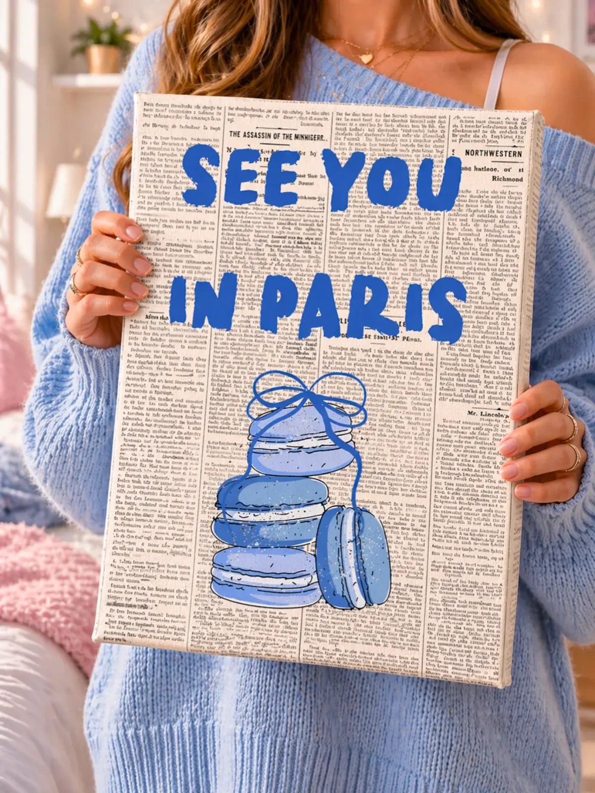 
A girl in a blue sweater holds the Doodles and Saffron See You In Paris canvas print with blue text and macarons over a newspaper print.