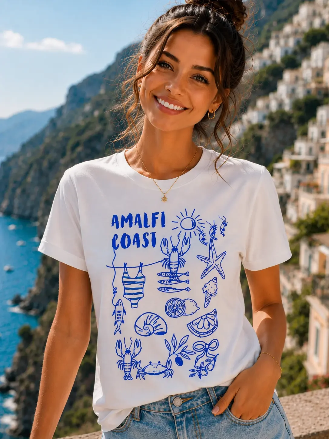 A smiling young woman wearing a white crewneck t-shirt featuring a blue Amalfi Coast icon graphic print, standing against a coastal backdrop. (Copy)