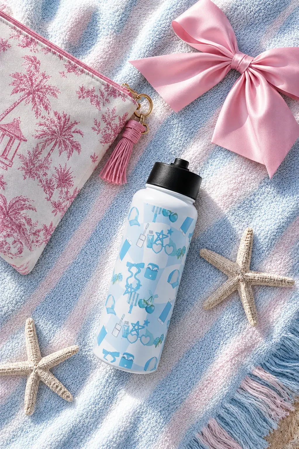 Flat lay of a blue doodle-print water bottle, pink toile bag, large satin bow, and starfishes on a striped towel.