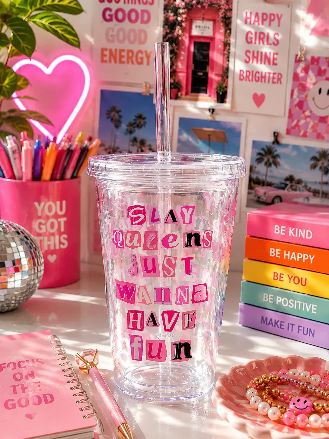 Hand holding a preppy "Slay Queens Just Wanna Have Fun" tumbler cup with iced coffee on an aesthetic pink desk setup.