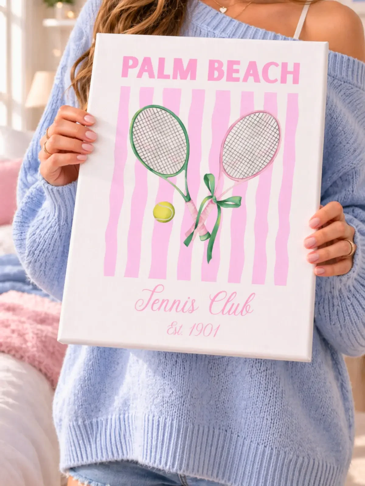 A young woman in a cozy light blue sweater holding a white rectangle wall art canvas printed with a pink-striped Palm Beach Tennis Club design. (Copy)