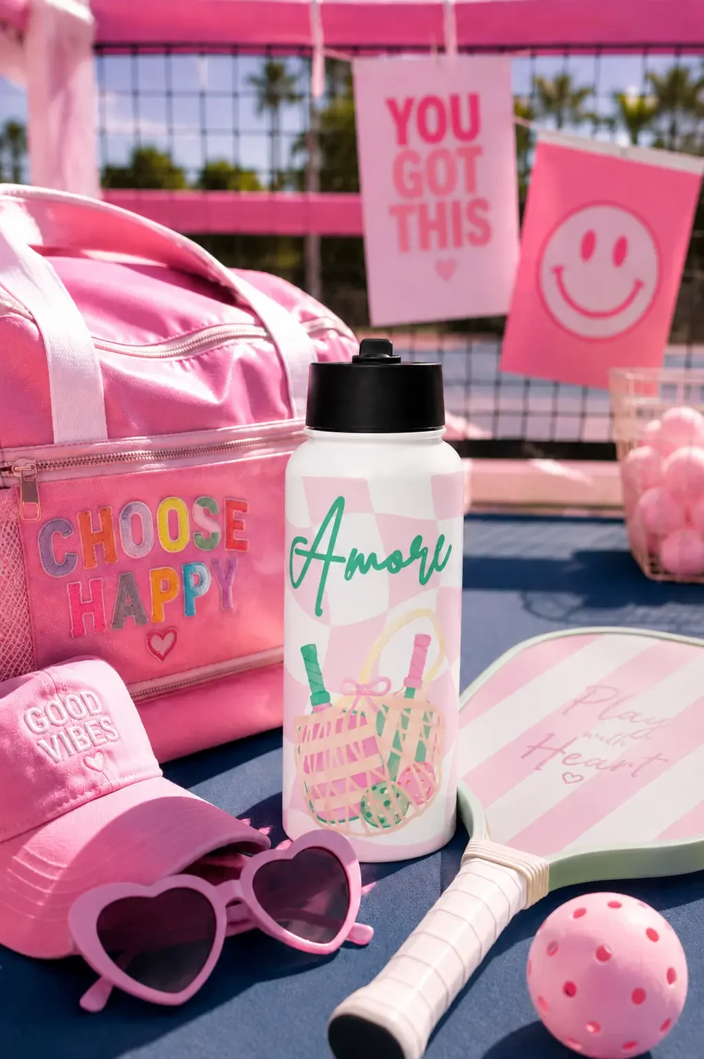 Flat lay of a pink Amore print water bottle, mint green pickleball paddles, stacked books, and pink sunglasses.