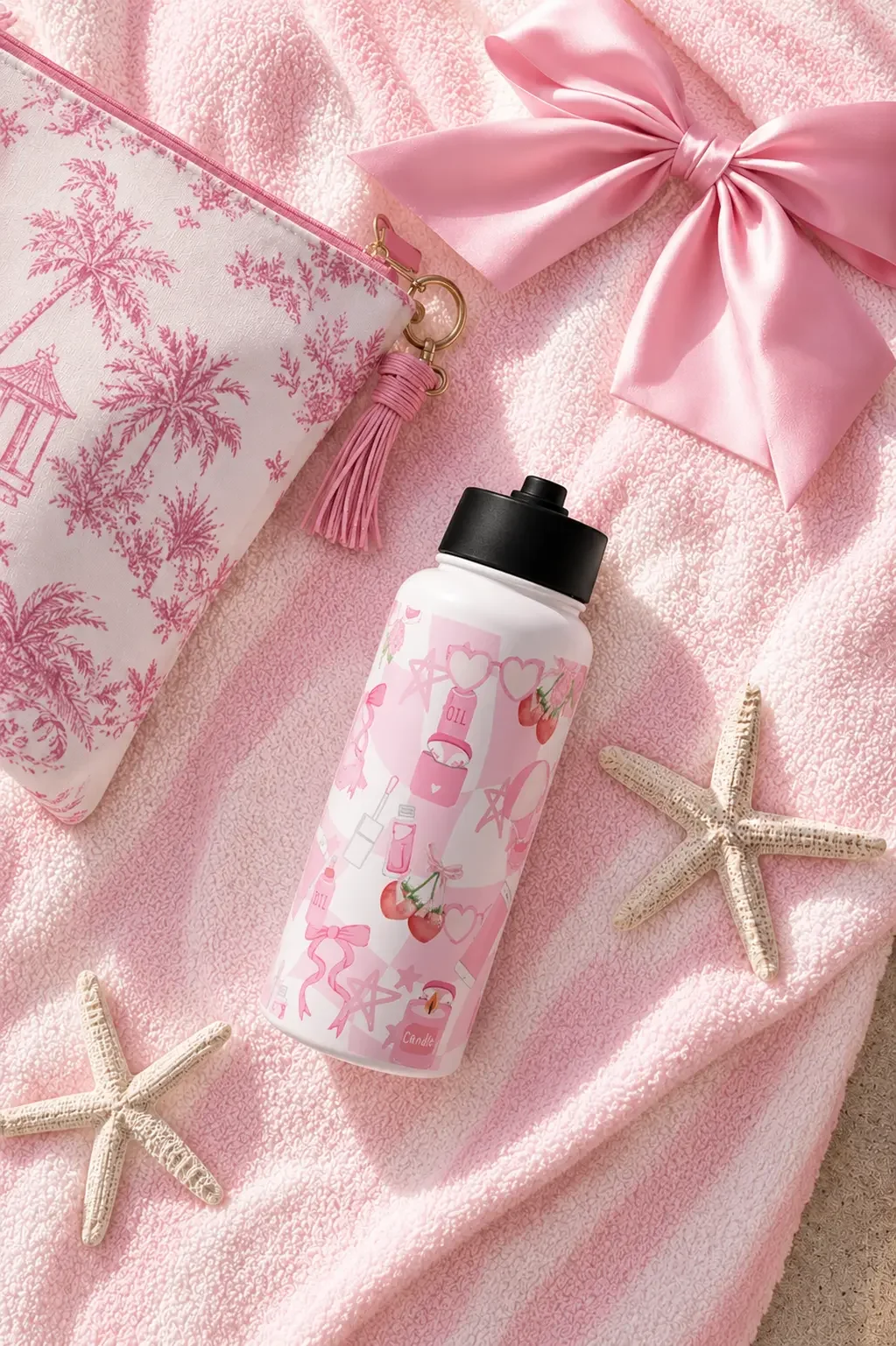 Flat lay of a pink cherry-print water bottle, toile makeup bag, large satin bow, and starfishes on a striped towel.