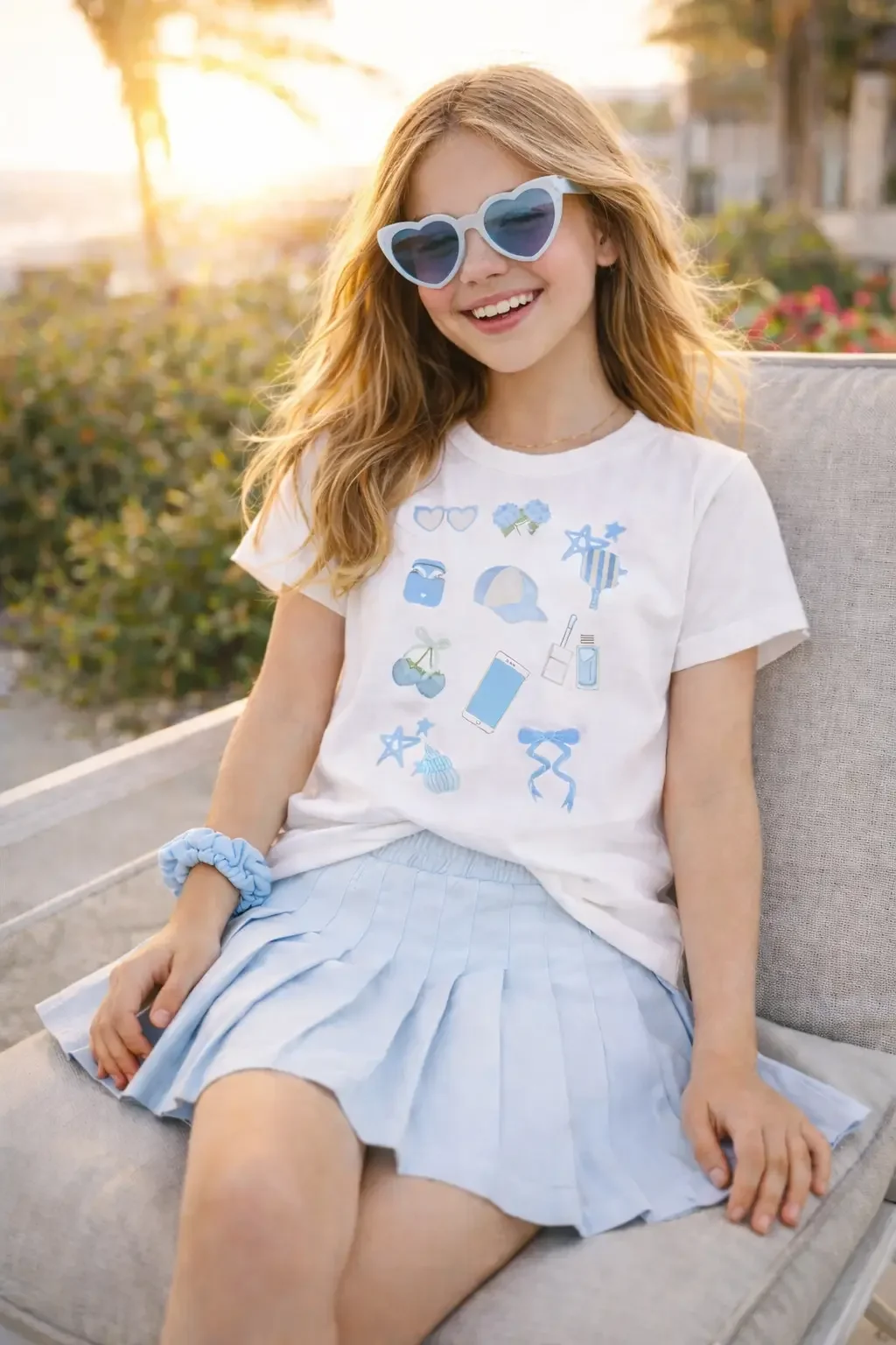 Young girl smiling on a beach lounge chair wearing a white preppy icon tee, light blue pleated skirt, and blue sunglasses.