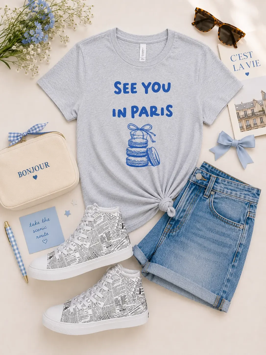 Flat lay display of a grey See You In Paris macaron graphic tee, denim shorts, newspaper-print sneakers, a Bonjour pouch, and sunglasses.