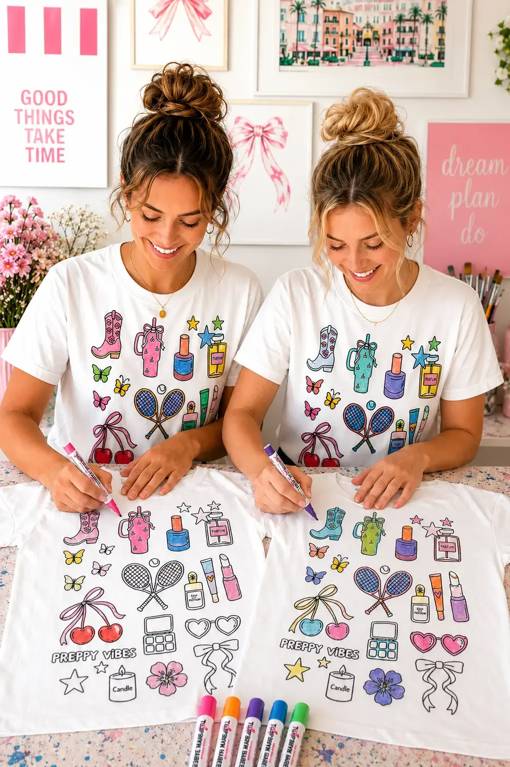 Two smiling young women with blonde and brunette top knot bun hairstyles sitting side by side at a table coloring matching preppy vibes doodle graphic t-shirts with colorful fabric markers.