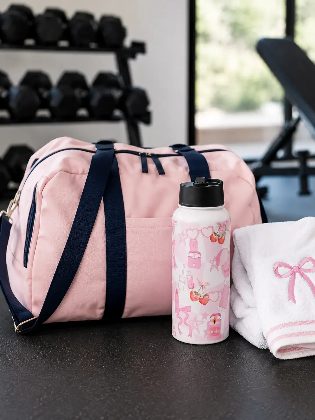 A pink graphic water bottle standing next to a pastel pink gym duffel bag and a white towel with a pink bow.