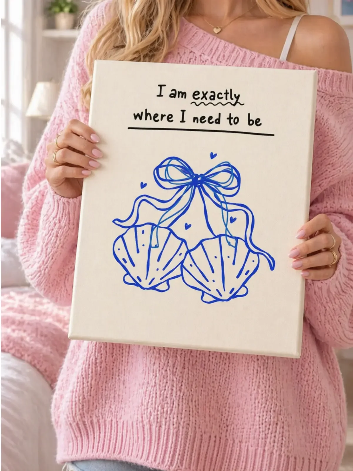 Model wearing a pink knit sweater holding the coastal blue seashell I Am Exactly Where I Need to Be canvas print by Doodles and Saffron.