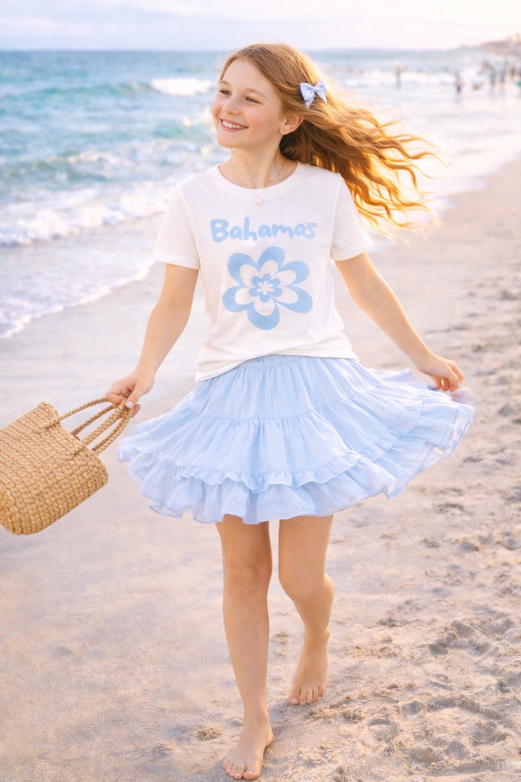 Girl twirling on beach in white Bahamas flower graphic tee and blue ruffle skirt.