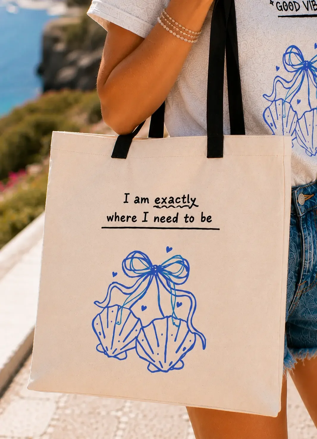 Close-up detail of a white tote bag with blue seashell bow graphic art and text reading I am exactly where I need to be.