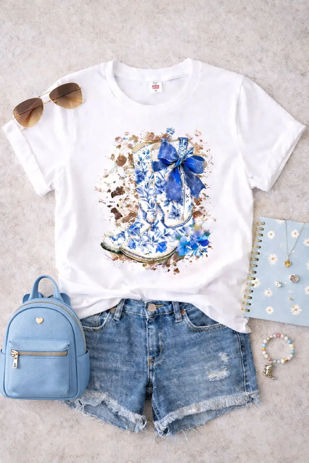 Flat lay of a white graphic tee featuring blue floral chinoiserie cowboy boots, styled with denim shorts and a blue mini backpack.