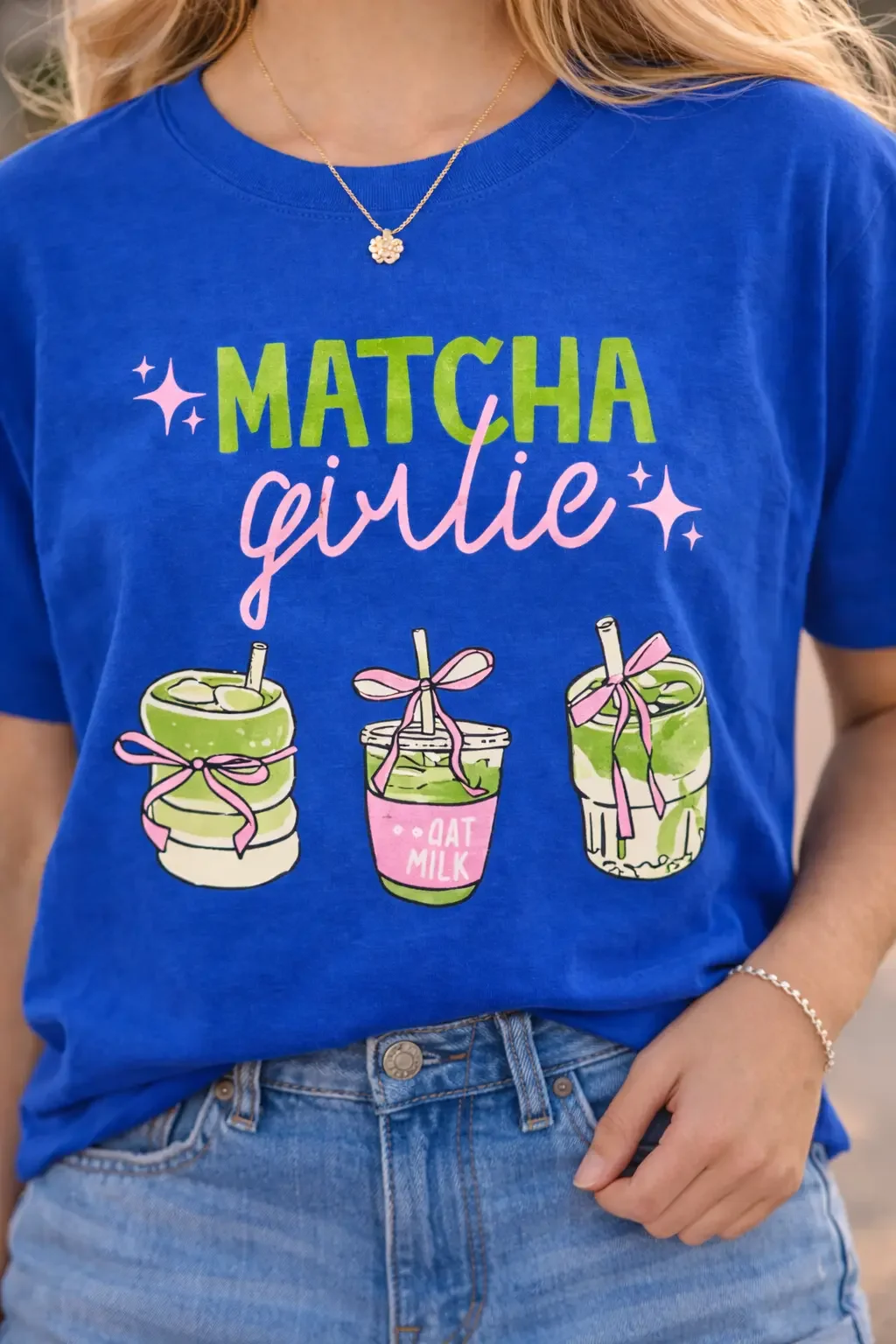 Close up of a blue graphic t-shirt featuring matcha girlie text with illustrations of iced matcha lattes tucked into denim jeans.