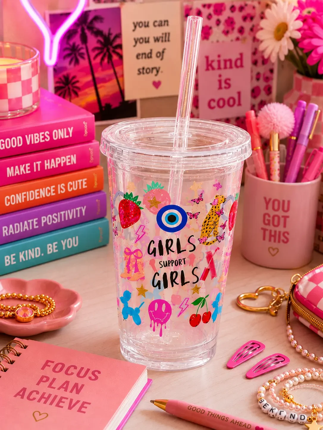 A clear preppy "Girls Support Girls" doodle tumbler cup on a pink desk surrounded by motivational books and hair accessories.