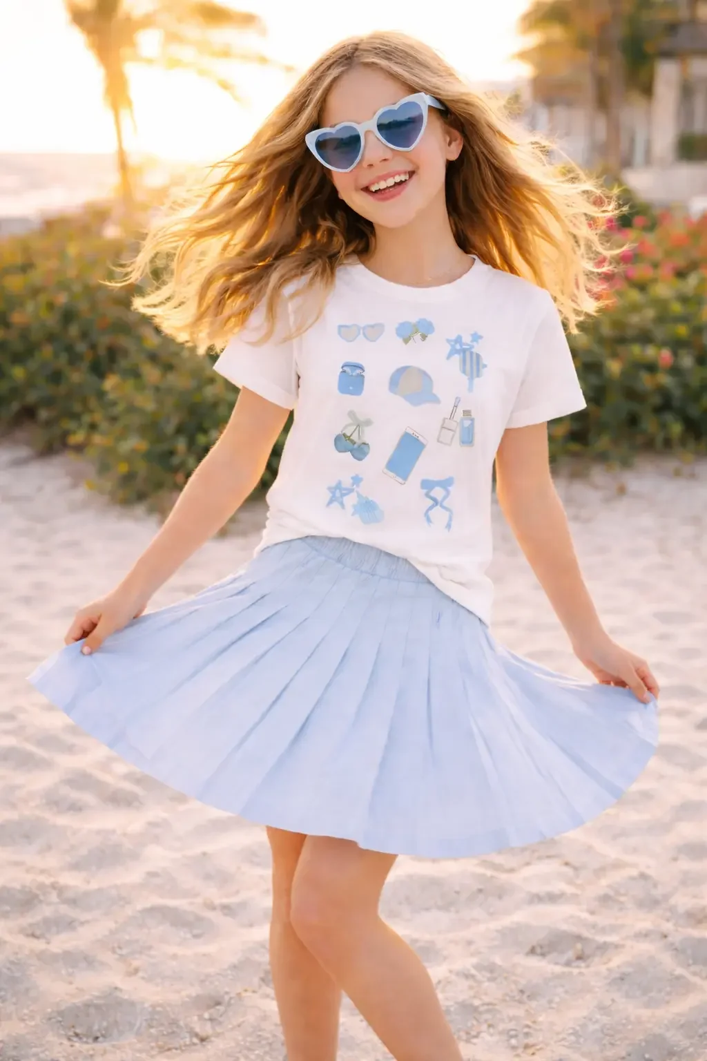 Young girl twirling on a beach wearing a white preppy icon t-shirt, blue pleated skirt, and heart-shaped sunglasses.