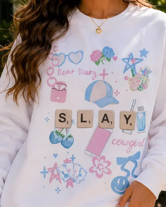 Close-up shot of a white crewneck sweatshirt displaying a colorful pastel graphic with Scrabble tiles spelling SLAY surrounded by doodles.