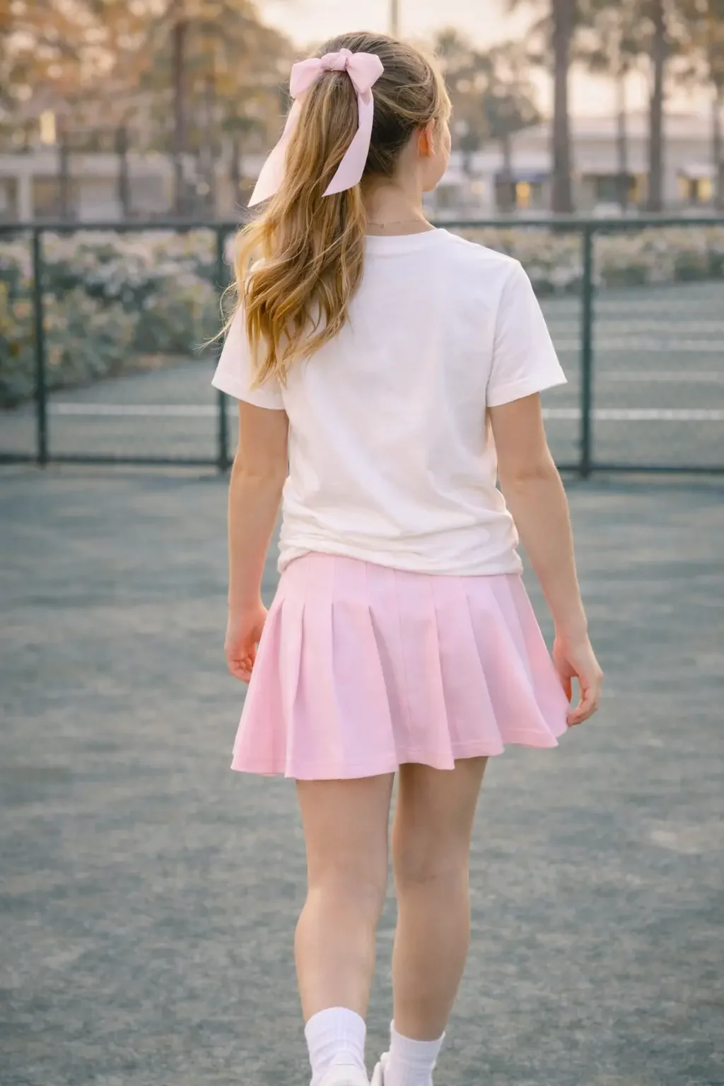 Back view of a girl standing on a tennis court wearing a white graphic t-shirt, a pink pleated tennis skirt, and a large pink satin hair bow clip.