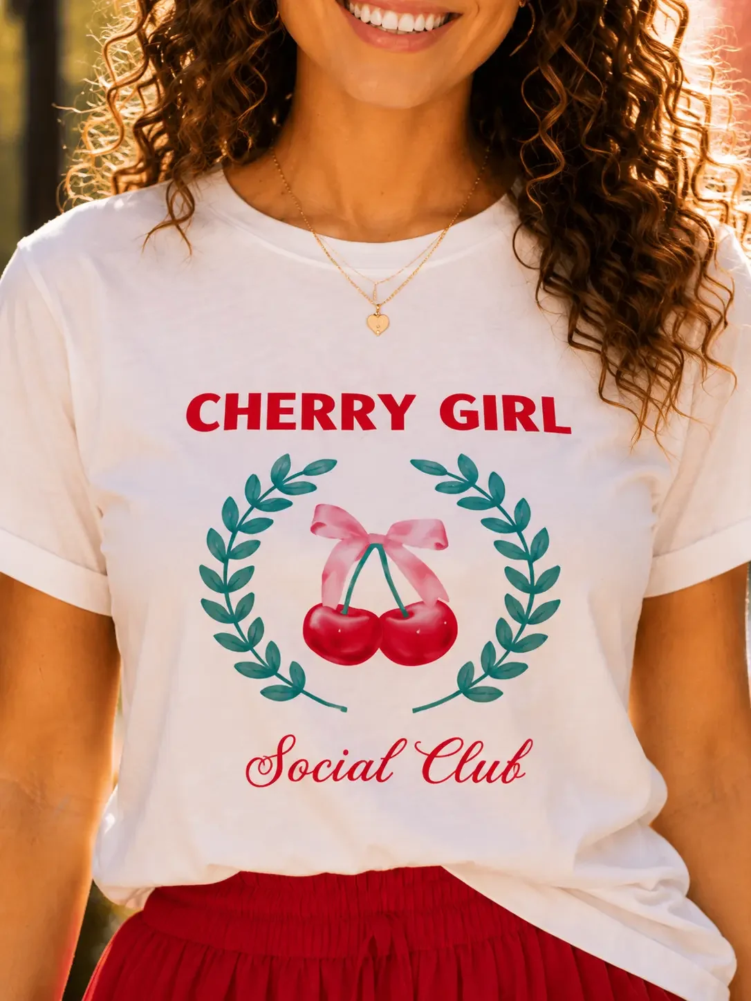 Close-up of a woman wearing a white "Cherry Girl Social Club" t-shirt with a cherry wreath graphic and gold necklace.