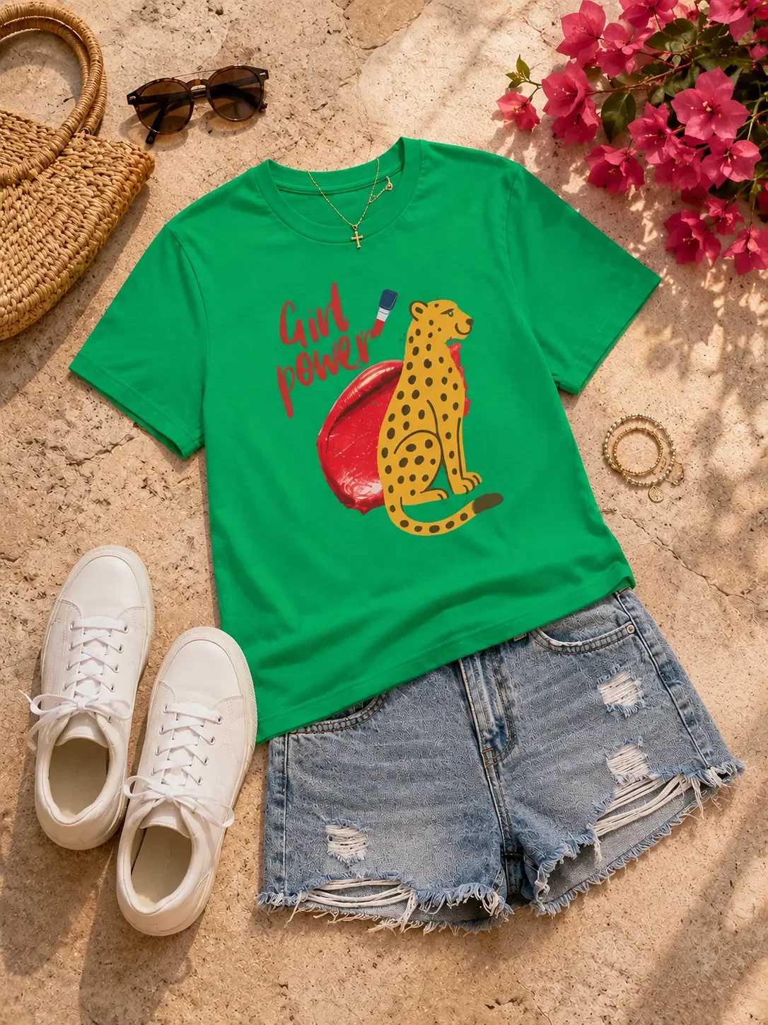 Flat lay display of a green Girl Power cheetah graphic t-shirt, distressed denim shorts, white sneakers, a straw tote bag, and sunglasses.