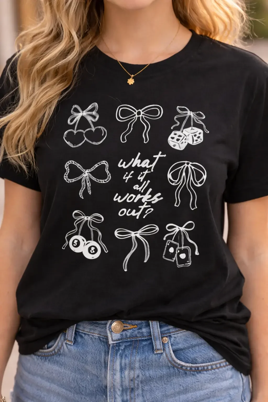 Close-up of a black graphic tee with white bow and cherry line art around the quote "what if it all works out?"