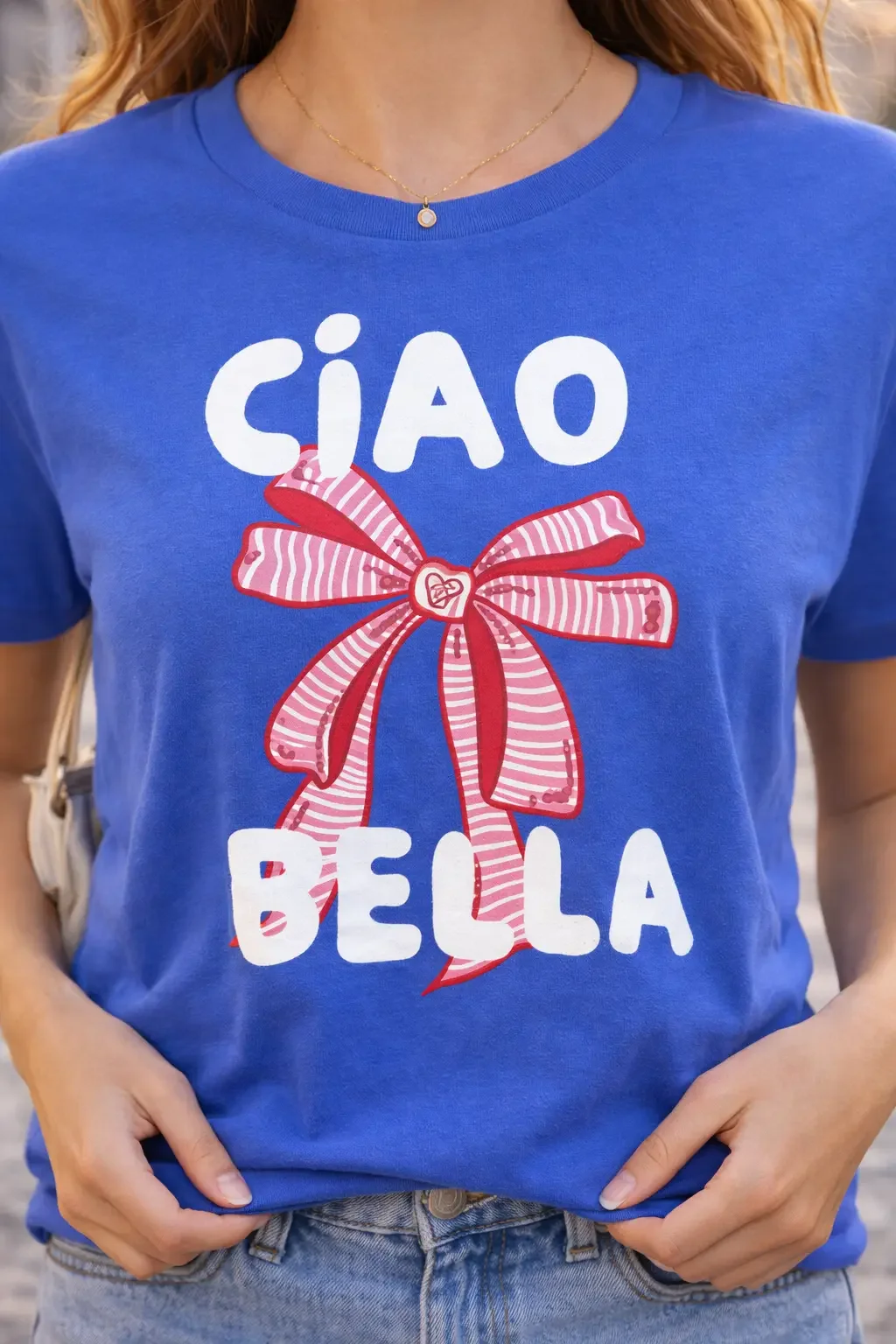 Close-up view of a blue crew neck t-shirt featuring the white text Ciao Bella and a large red-and-white striped ribbon bow graphic.