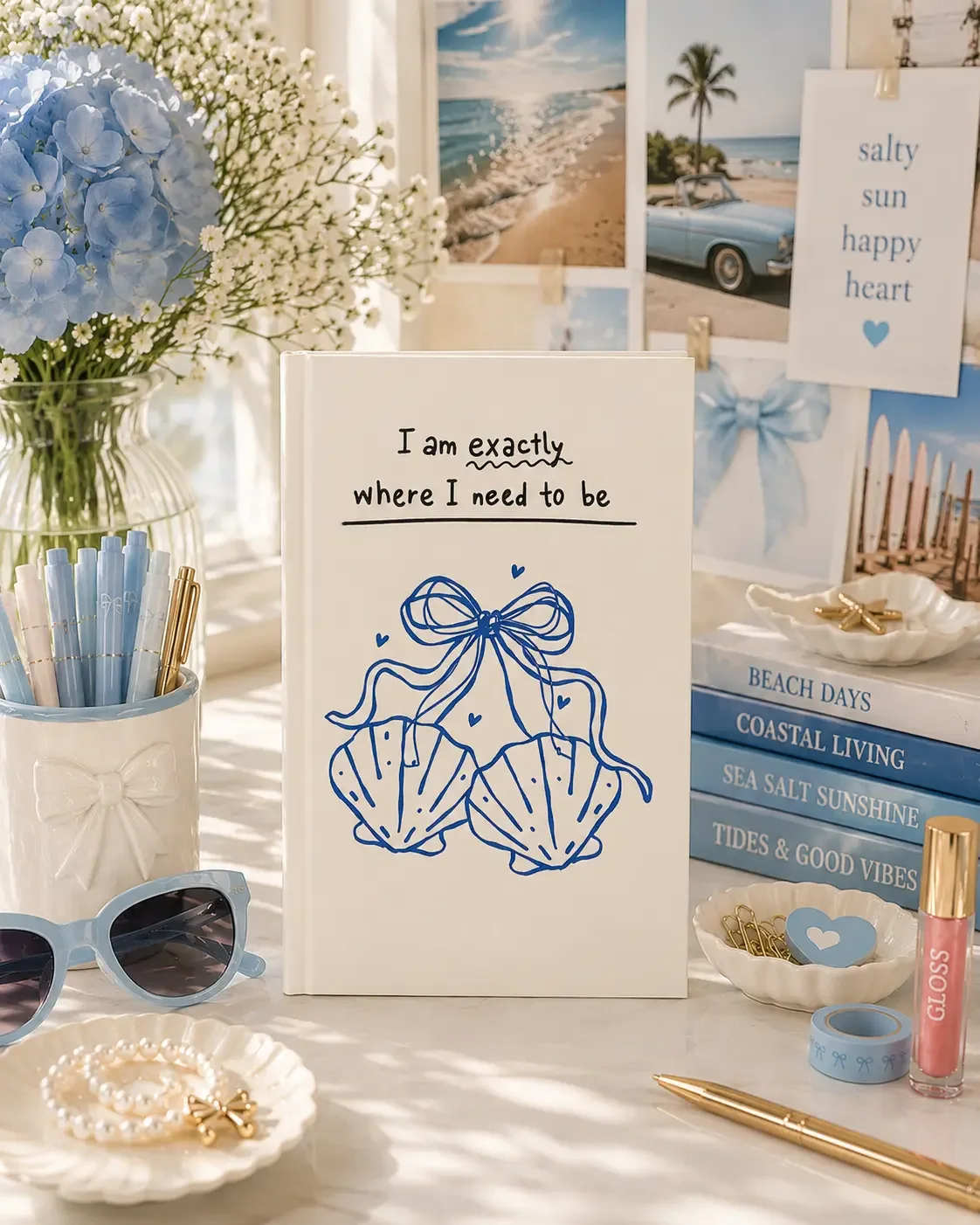 Coastal aesthetic desk flat lay featuring an "I am exactly where I need to be" seashell journal, blue sunglasses, stack of books, and hydrangeas.