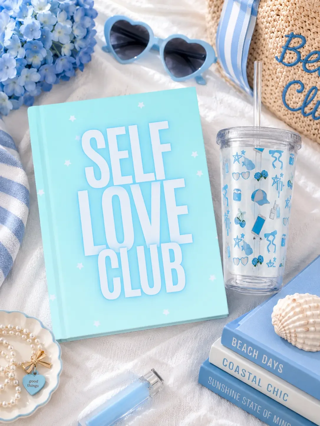 Flat lay of a mint green "Self Love Club" notebook, matching pattern tumbler, blue heart sunglasses, and decorative books on a beach towel.