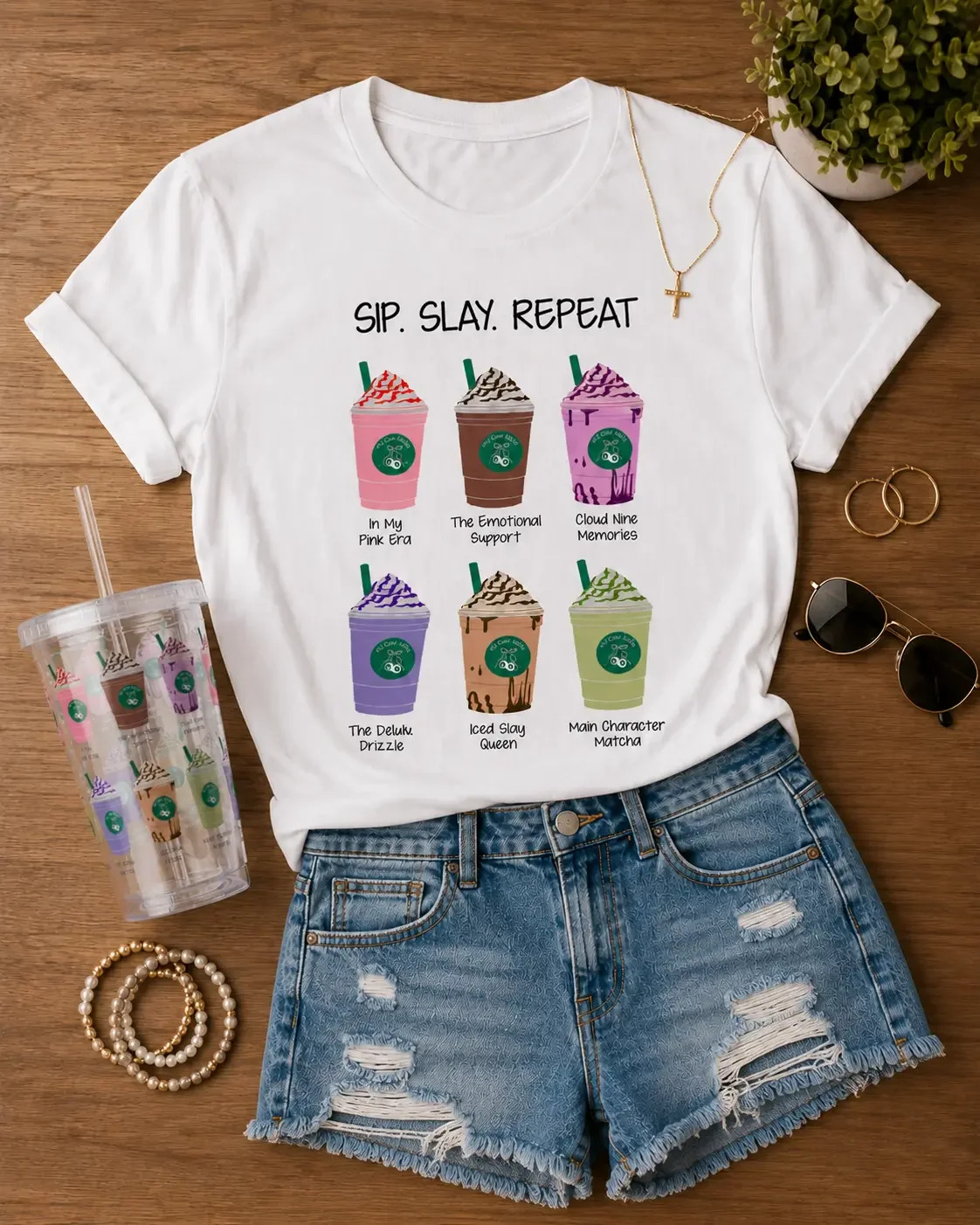 Flat lay arrangement of a white "Sip. Slay. Repeat." coffee t-shirt, distressed denim shorts, a tumbler, and jewelry.