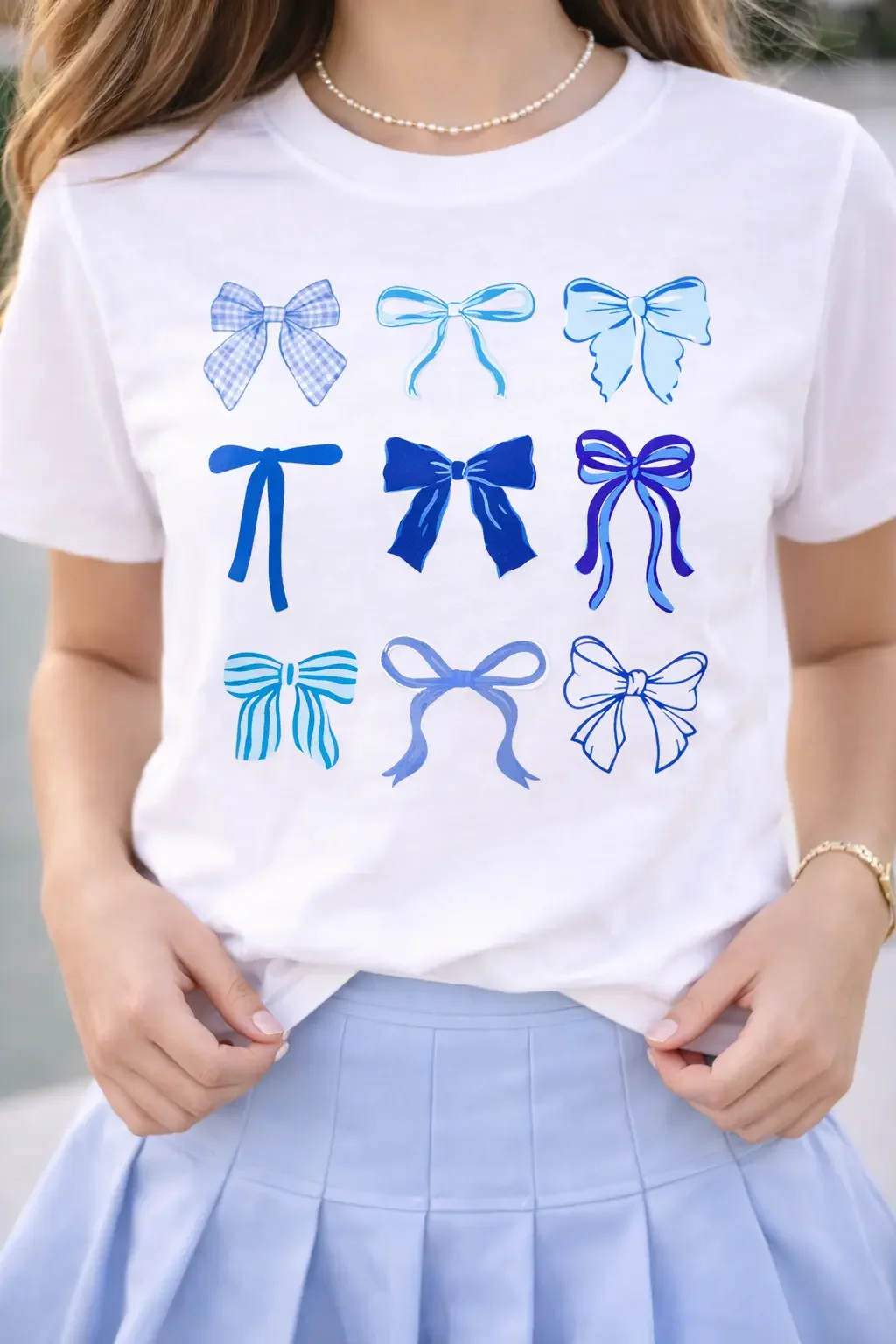 Close-up of a white graphic t-shirt featuring nine different blue ribbon bow designs, styled with a pleated skirt.