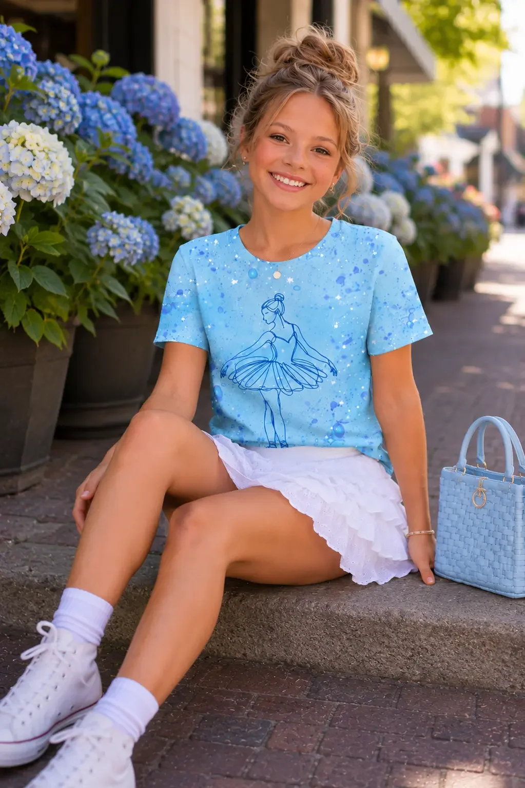 Smiling girl sitting on a concrete ledge next to blue hydrangeas wearing a blue paint-splatter ballerina graphic t-shirt, white tiered mini skirt, and white high-top sneakers.