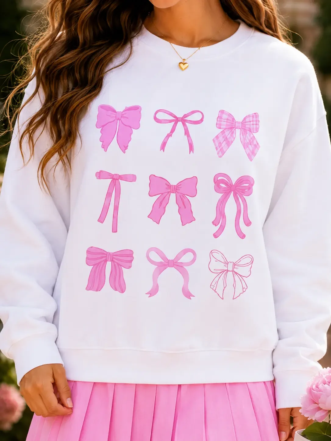 Close-up shot of a white crewneck sweatshirt featuring a central graphic print displaying a nine-piece grid of various pink ribbon bows.
