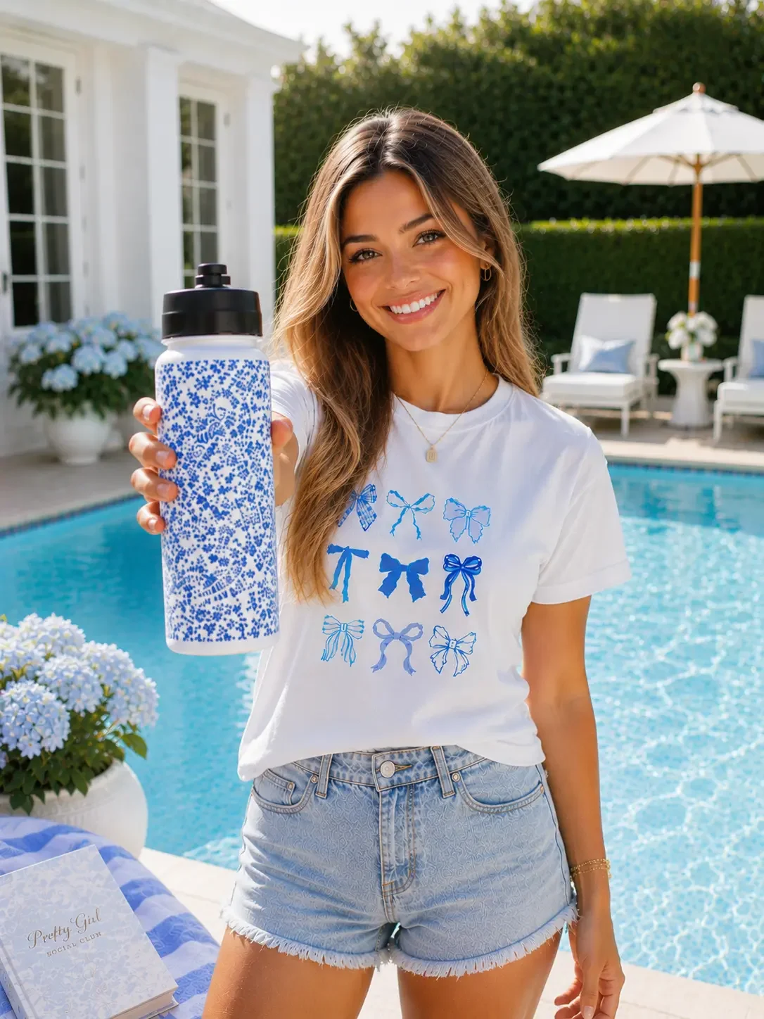 A woman holds a blue floral water bottle poolside, wearing denim shorts and a white t-shirt featuring blue bow designs.