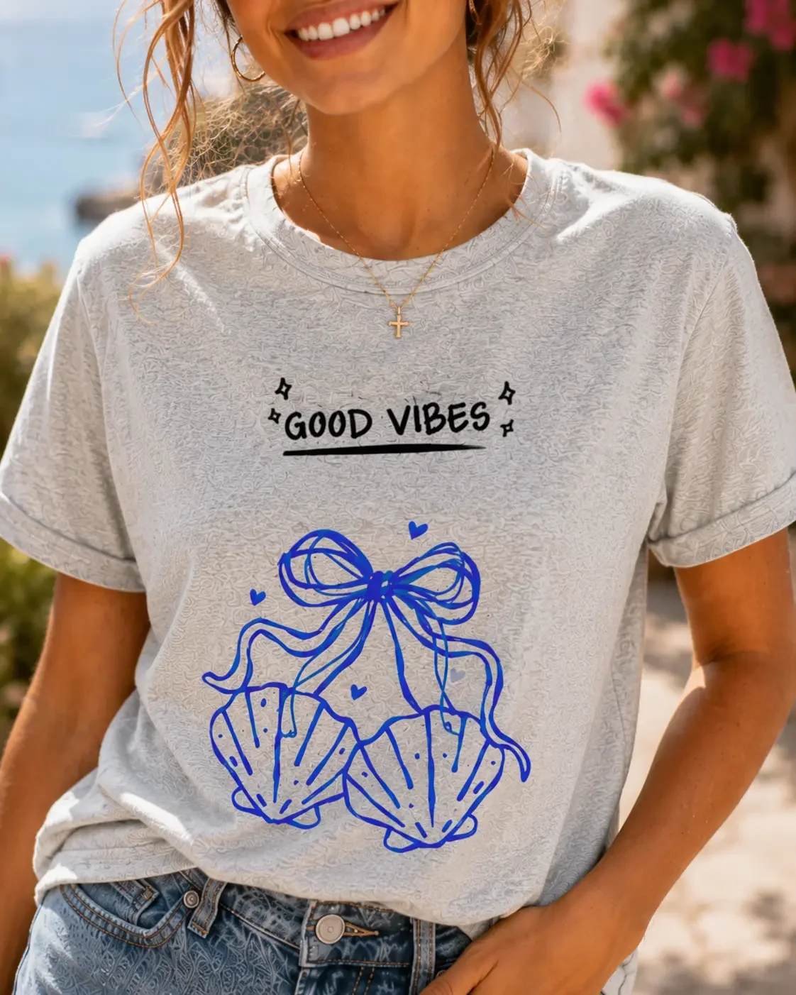Close-up view of a grey crew neck t-shirt featuring black Good Vibes text above a blue seashell and ribbon bow graphic illustration.