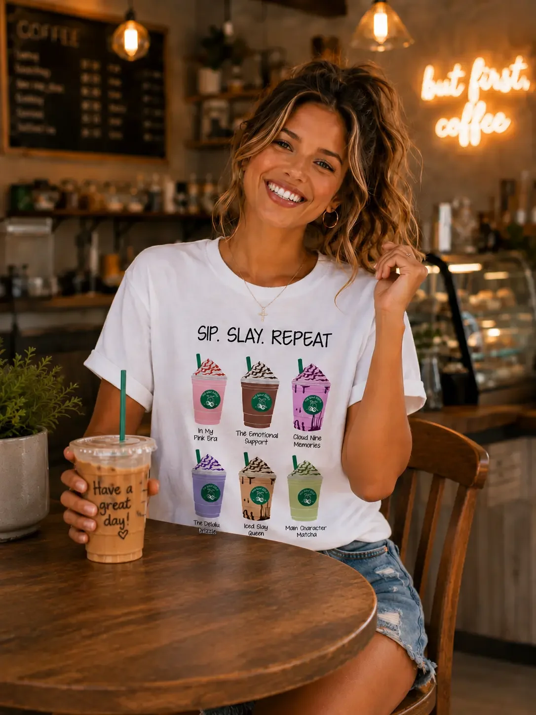 Smiling woman sitting at a table with an iced coffee, wearing a white "Sip. Slay. Repeat." graphic t-shirt in a cafe.