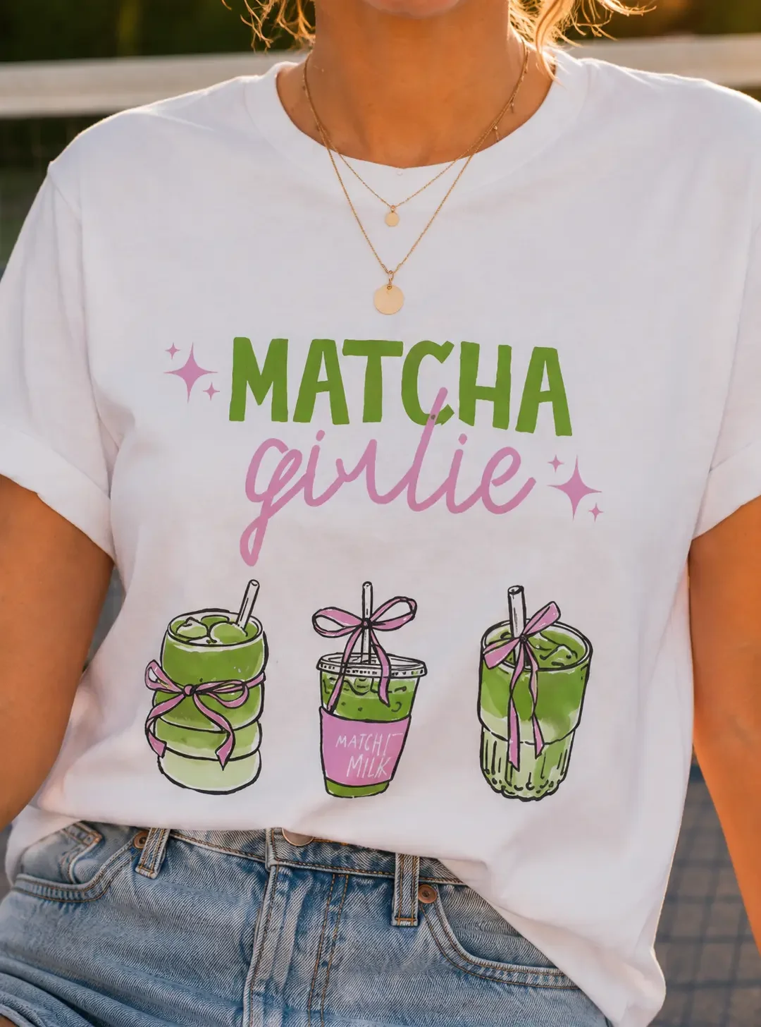 Close-up view of a white crew neck t-shirt featuring Matcha Girlie text above three distinct iced matcha beverage graphic illustrations.
