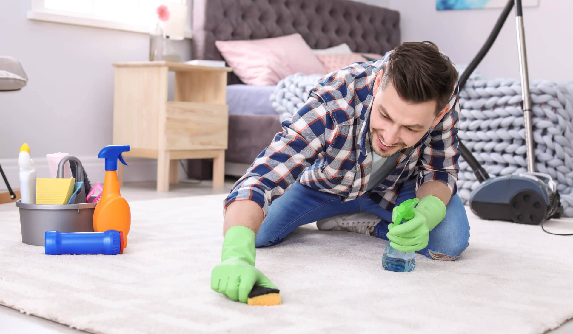 Man cleaning carpet with sponge and spray bottle in a living room, wearing green gloves, with cleaning supplies and a vacuum cleaner in the background.