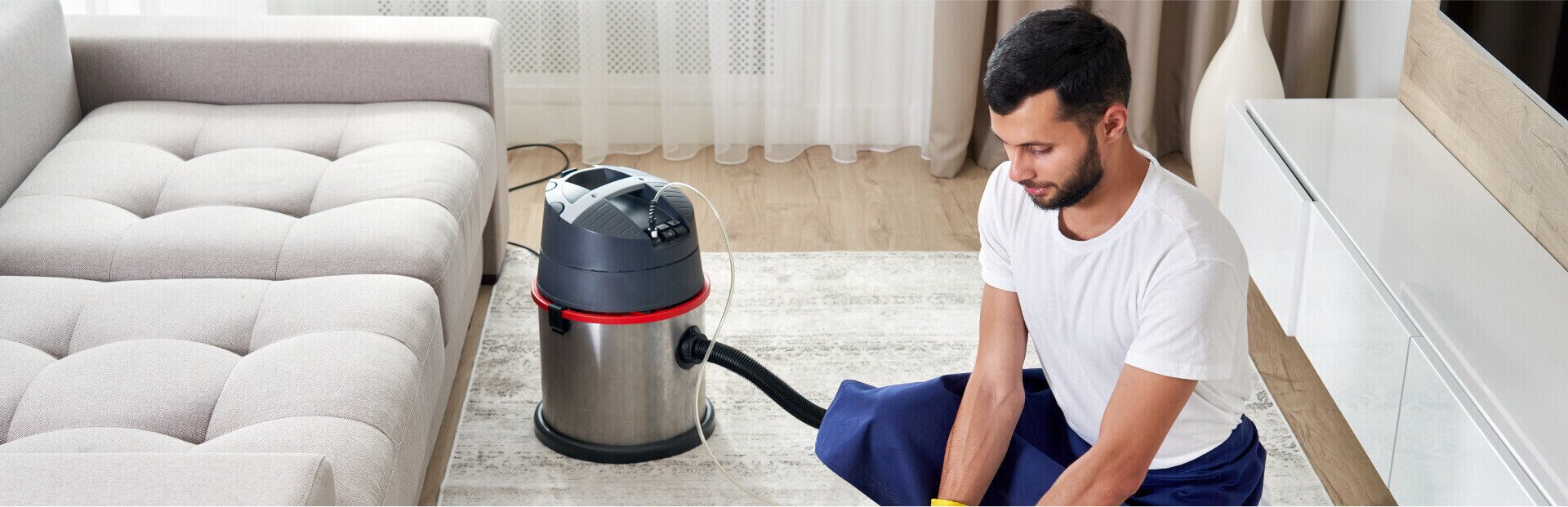 Man cleaning carpet with vacuum in living room next to sofa and entertainment unit.