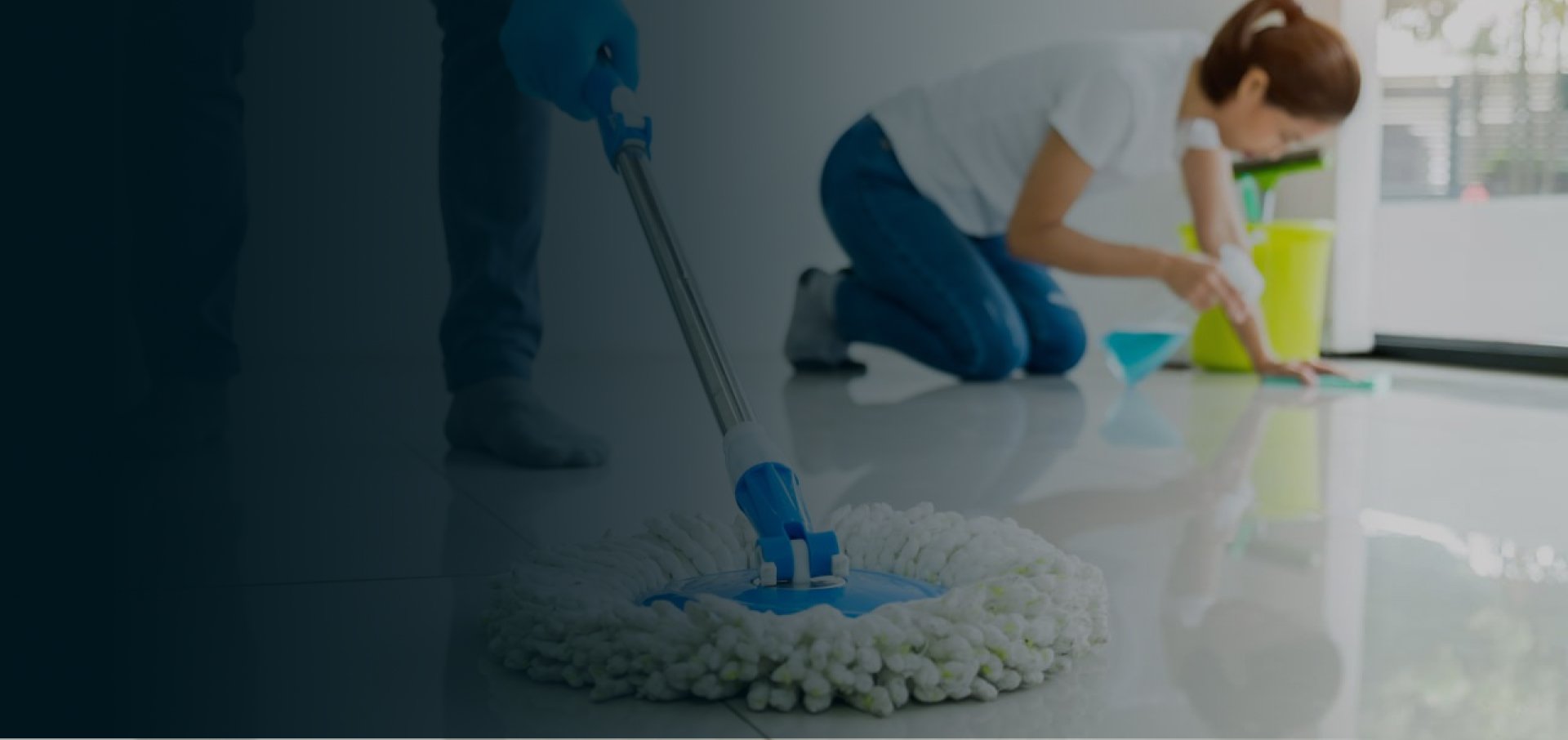 Person cleaning floor with a mop in the foreground, another person scrubbing with a cloth in the background, and cleaning supplies nearby.