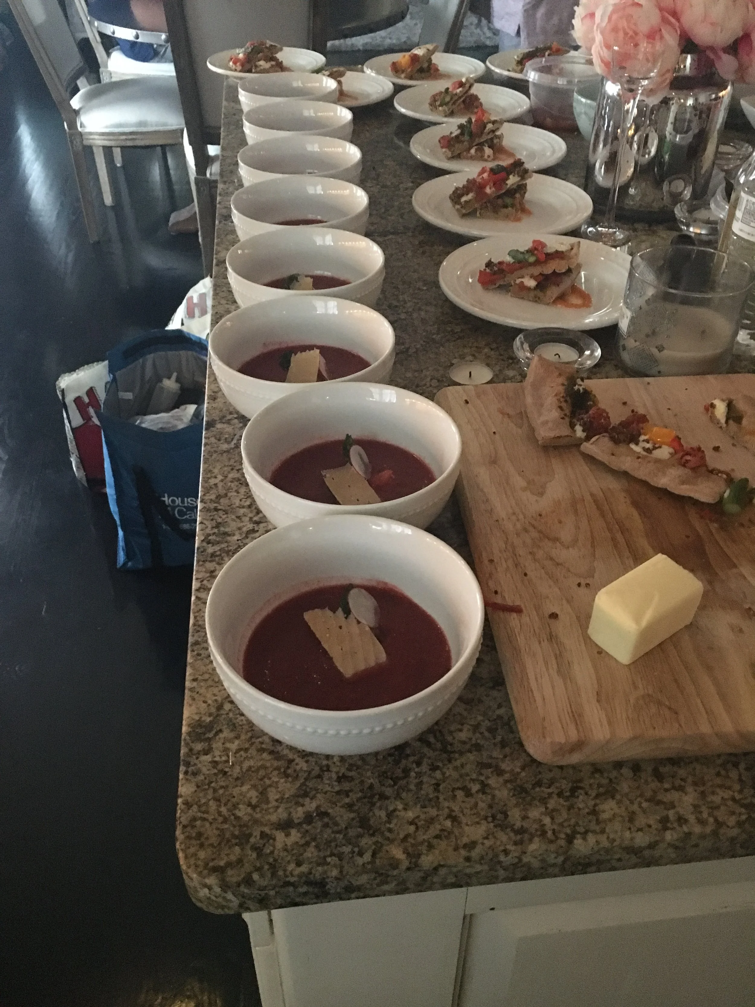 Several bowls of what appears to be a berry soup or puree with a piece of white chocolate or cheese on top, set on a kitchen counter. In the background, there are plates with food, a cutting board with a half-eaten pizza crust, and some decorative items like candles and flowers.