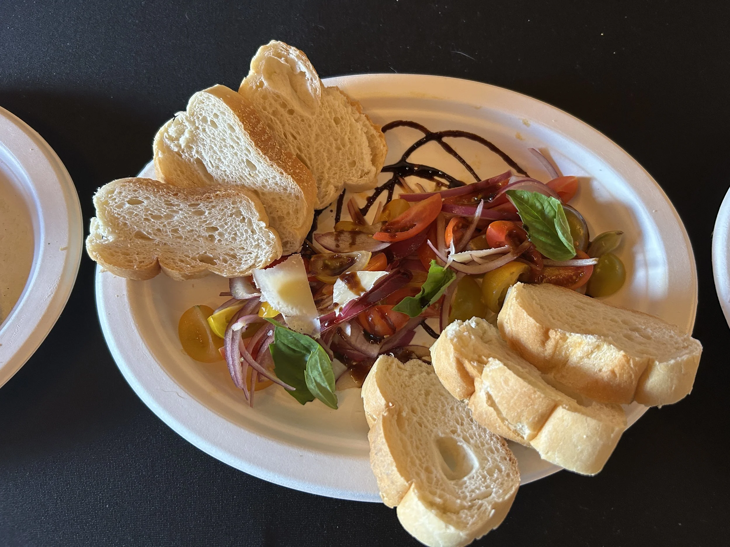 A white oval plate with sliced baguette bread, a small salad with cherry tomatoes, red onion, and basil leaves, drizzled with balsamic vinegar, on a black surface.