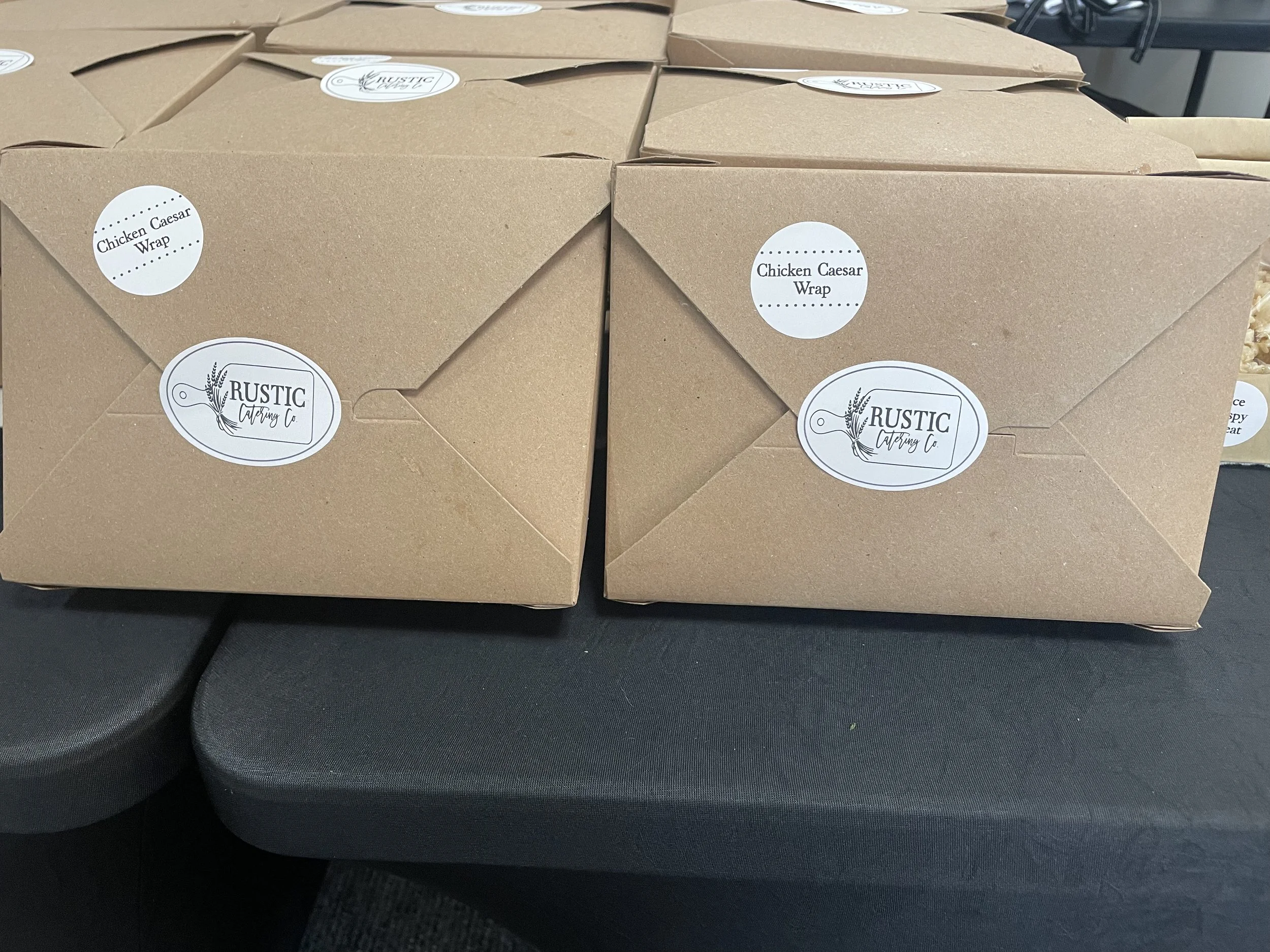 Multiple brown boxes labeled 'Chicken Caesar Wrap' and 'Rustic Catering Co.' are stacked on a black surface.
