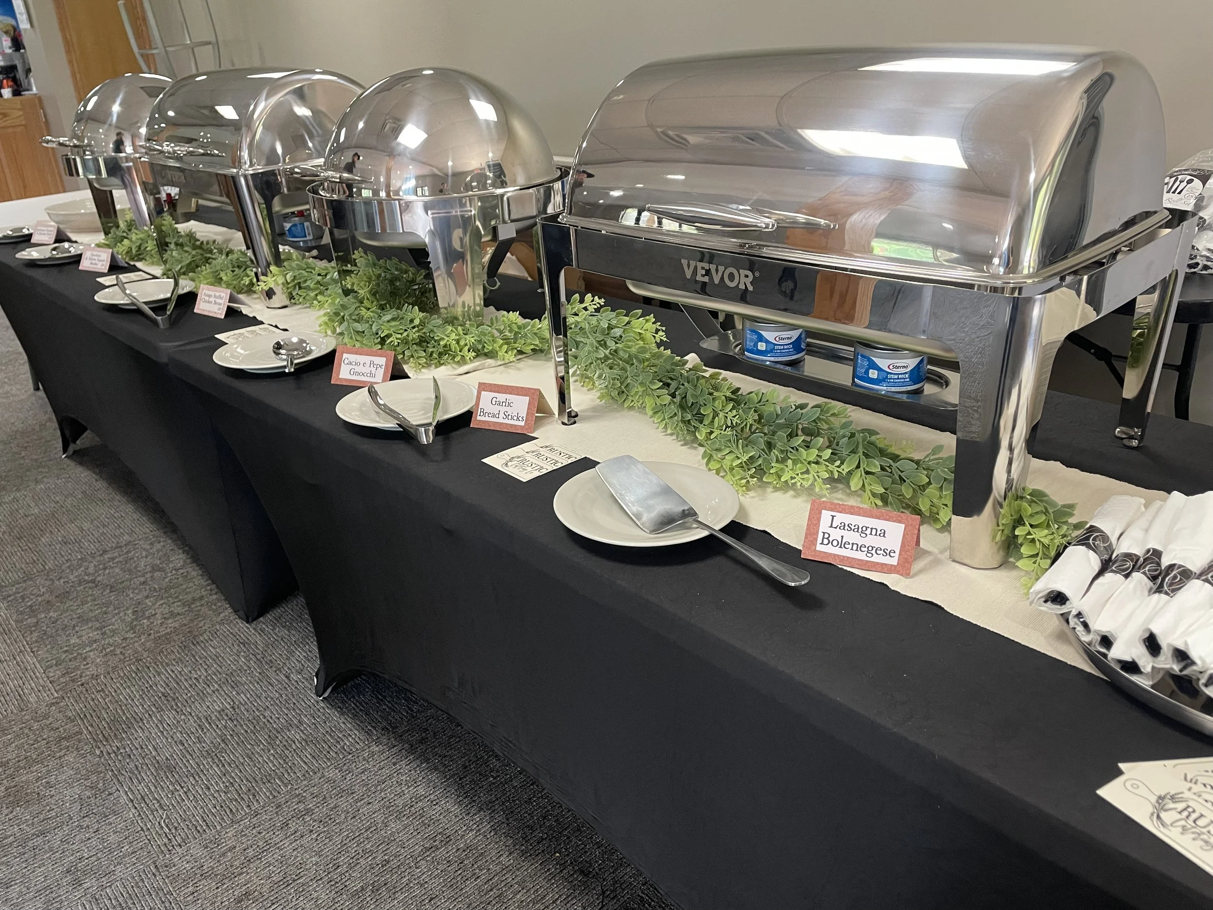 A buffet table with four metal chafing dishes, small plates, tongs, and various labels including "Gnocchi" and "Lasagna". Green foliage decorates the table.