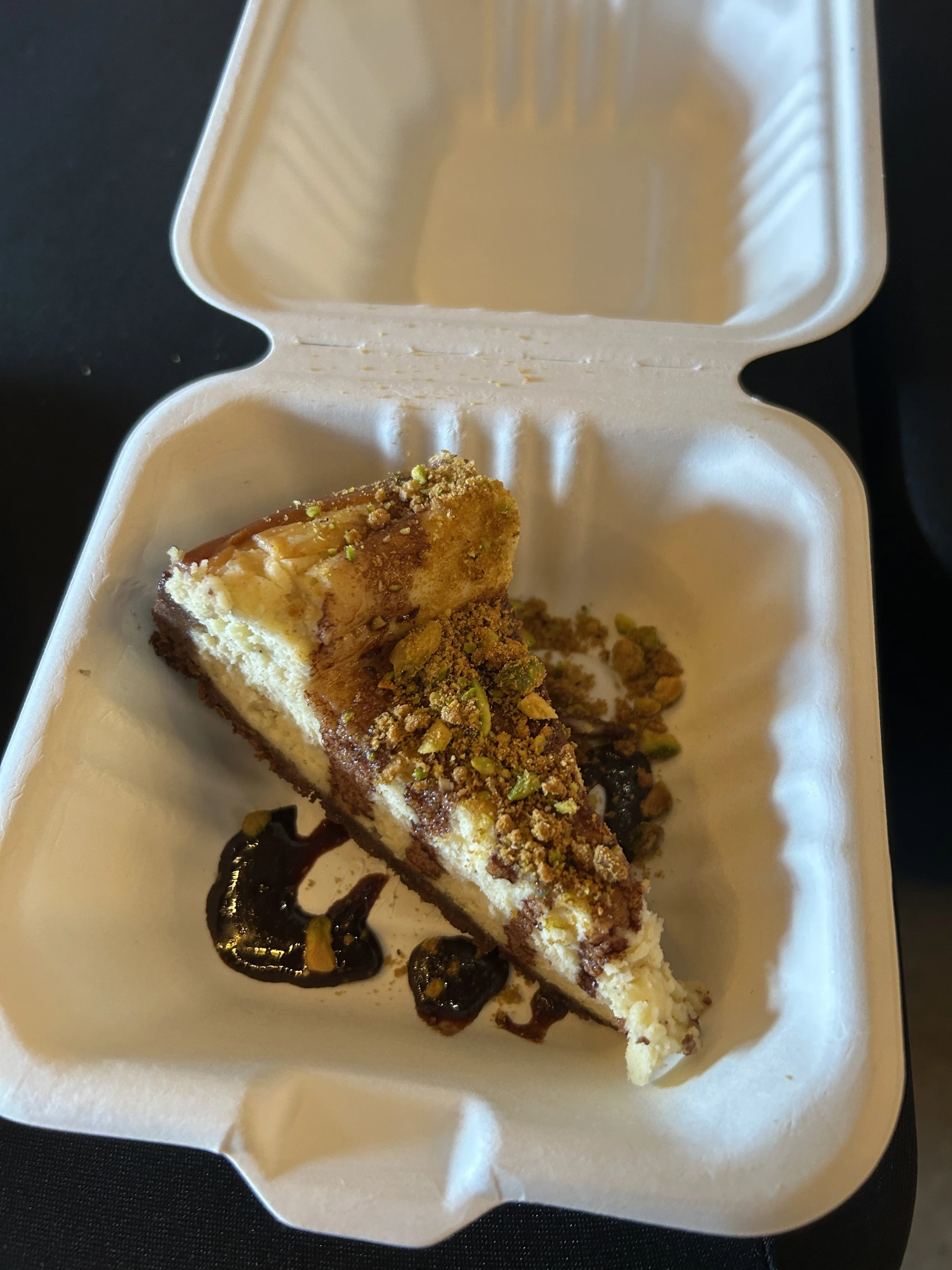 Slice of cheesecake with caramel drizzle, crushed nuts, and chocolate sauce on a white foam plate.