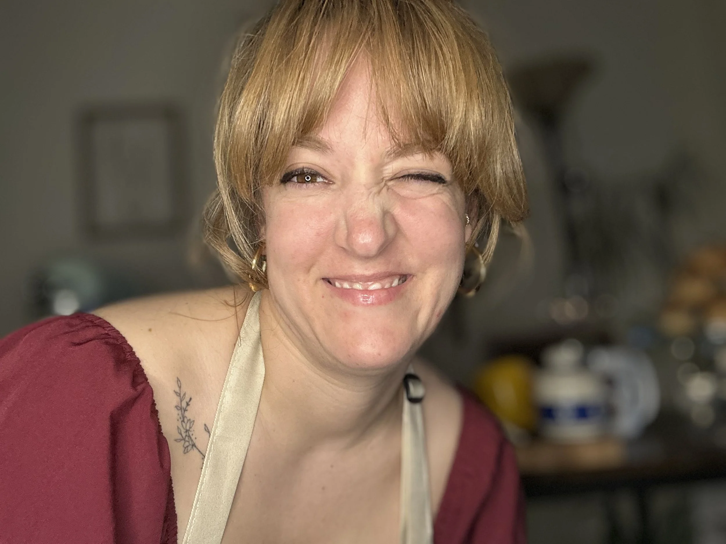 A smiling woman with short red hair, one eye closed and the other open, wearing a maroon top and a beige apron, standing in a kitchen.