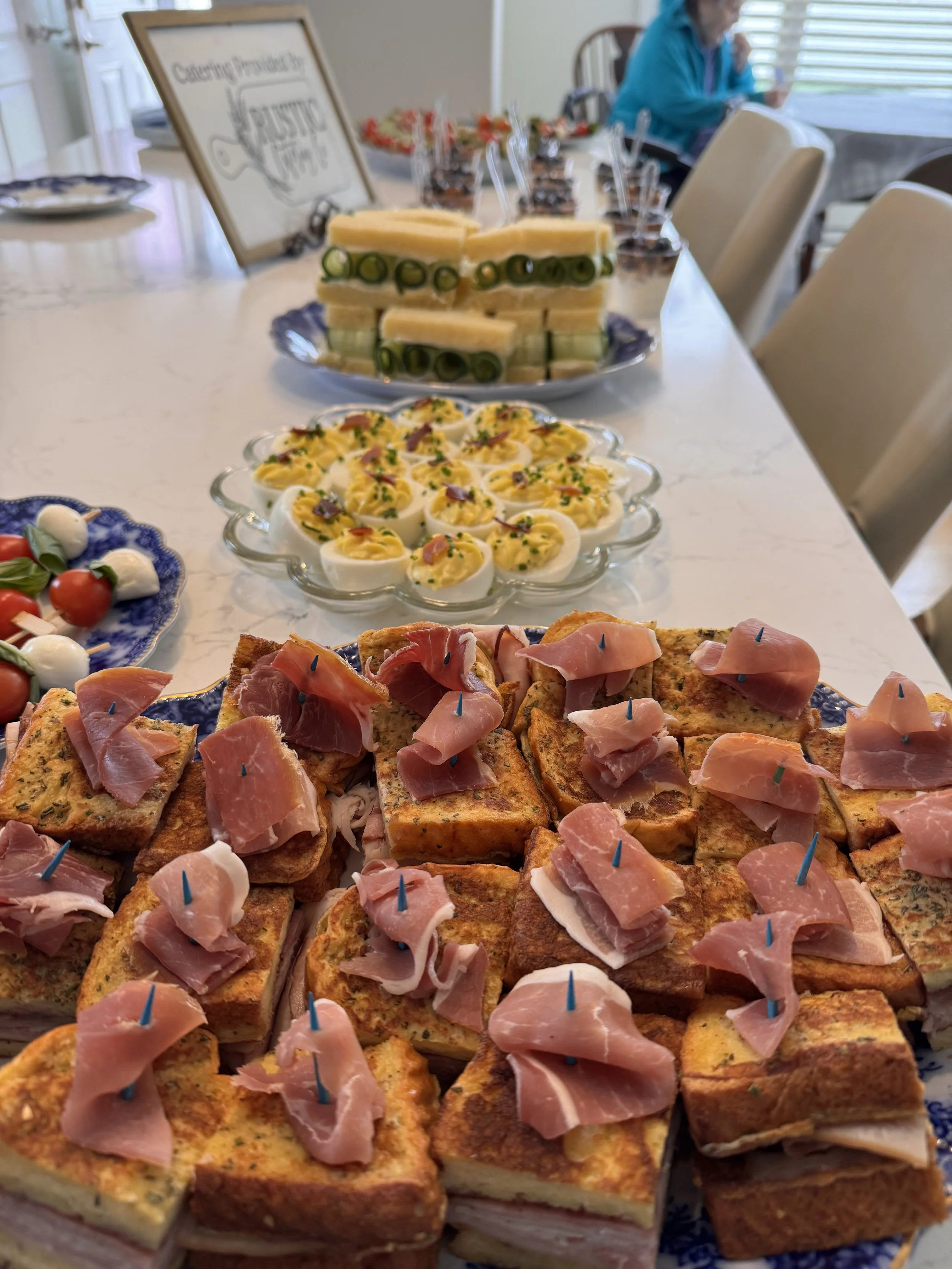 Plate of assorted finger foods, including mini sandwiches with cucumber and cheese, and slices of toasted bread with ham, on a table at a gathering.